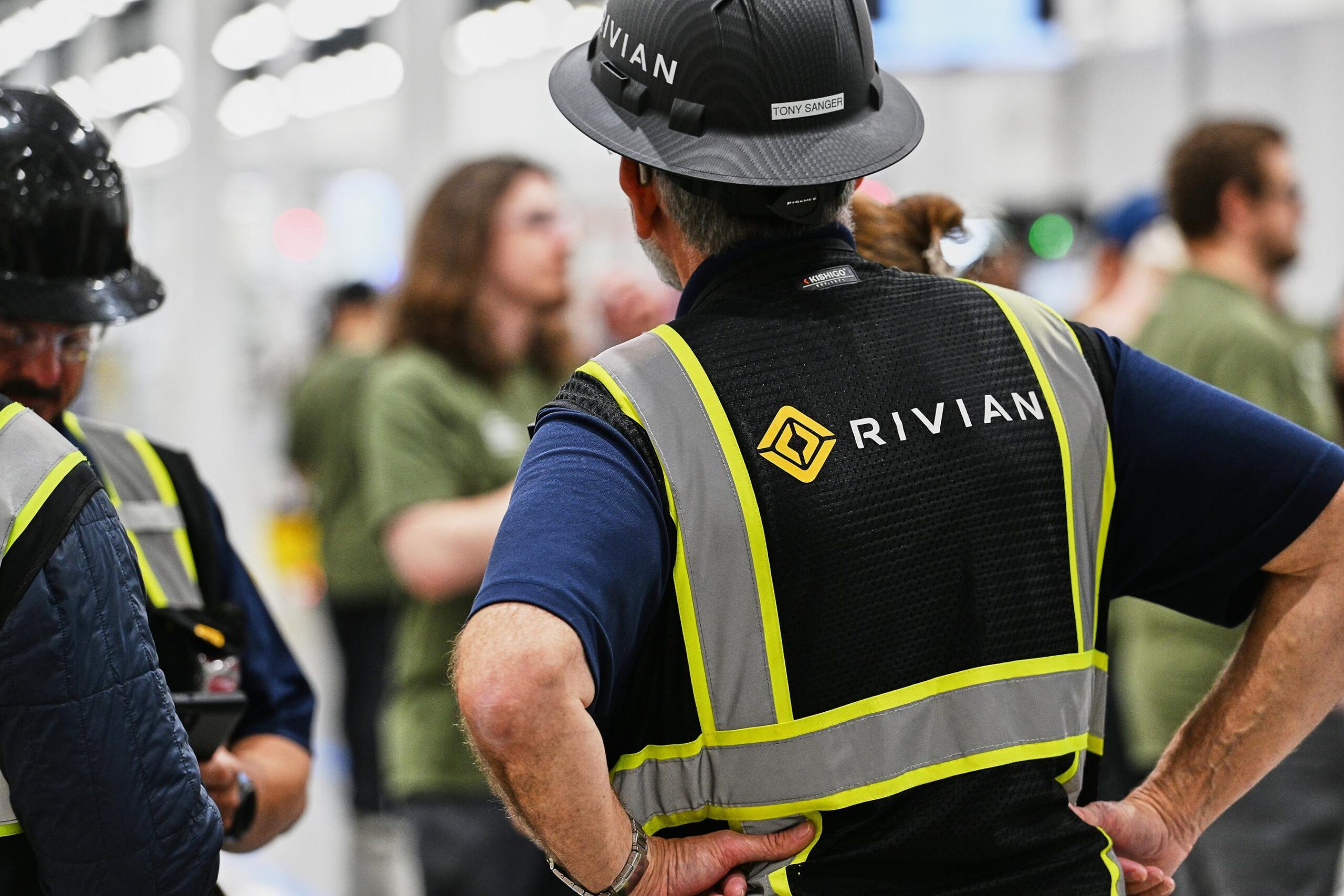 A person wears a black and yellow reflective vest with Rivian written on the back. A person wears a black and yellow reflective vest with Rivian written on the back.