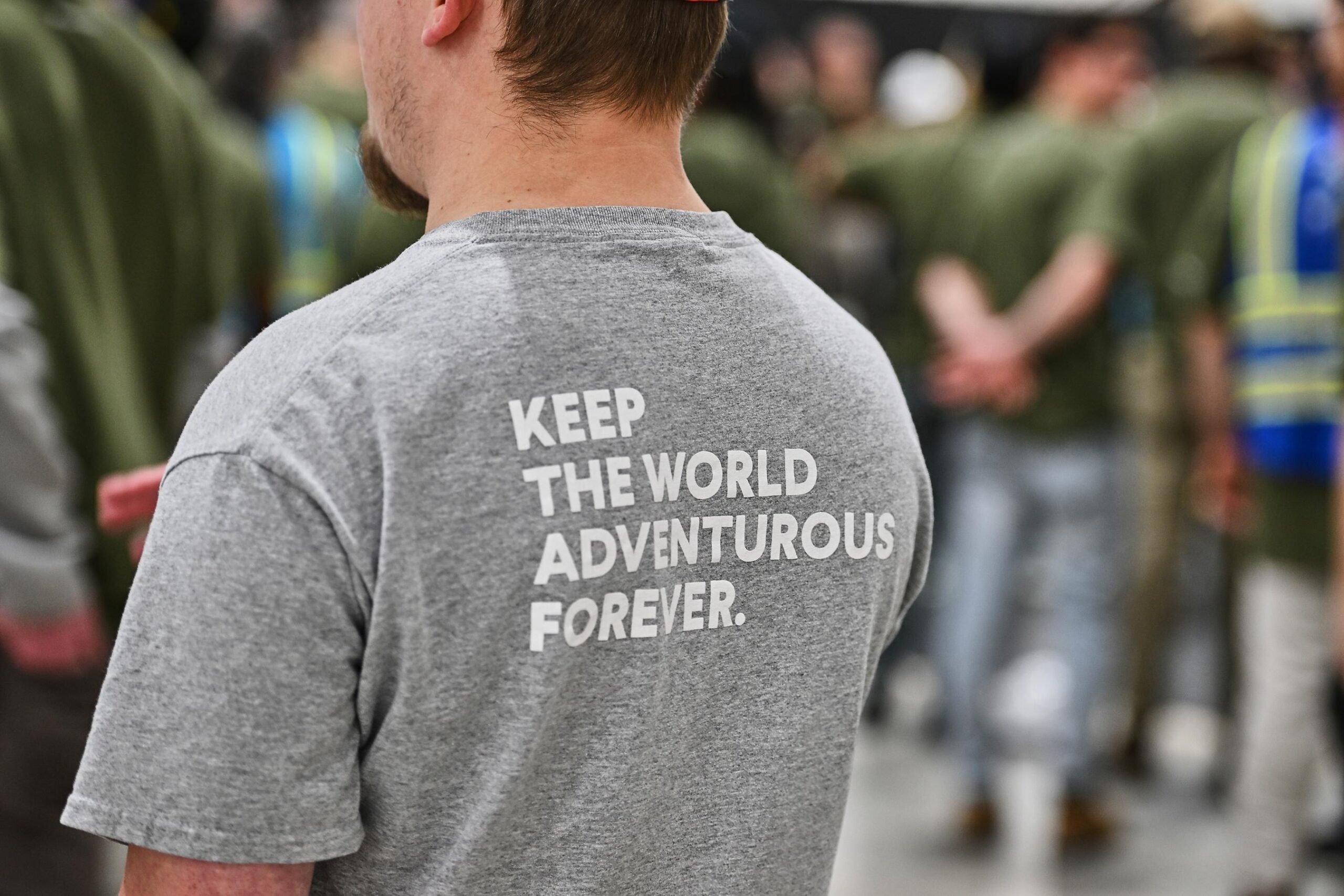A person with short hair wears a gray shirt that reads "keep the world adventurous forever" on the back. A person with short hair wears a gray shirt that reads "keep the world adventurous forever" on the back.