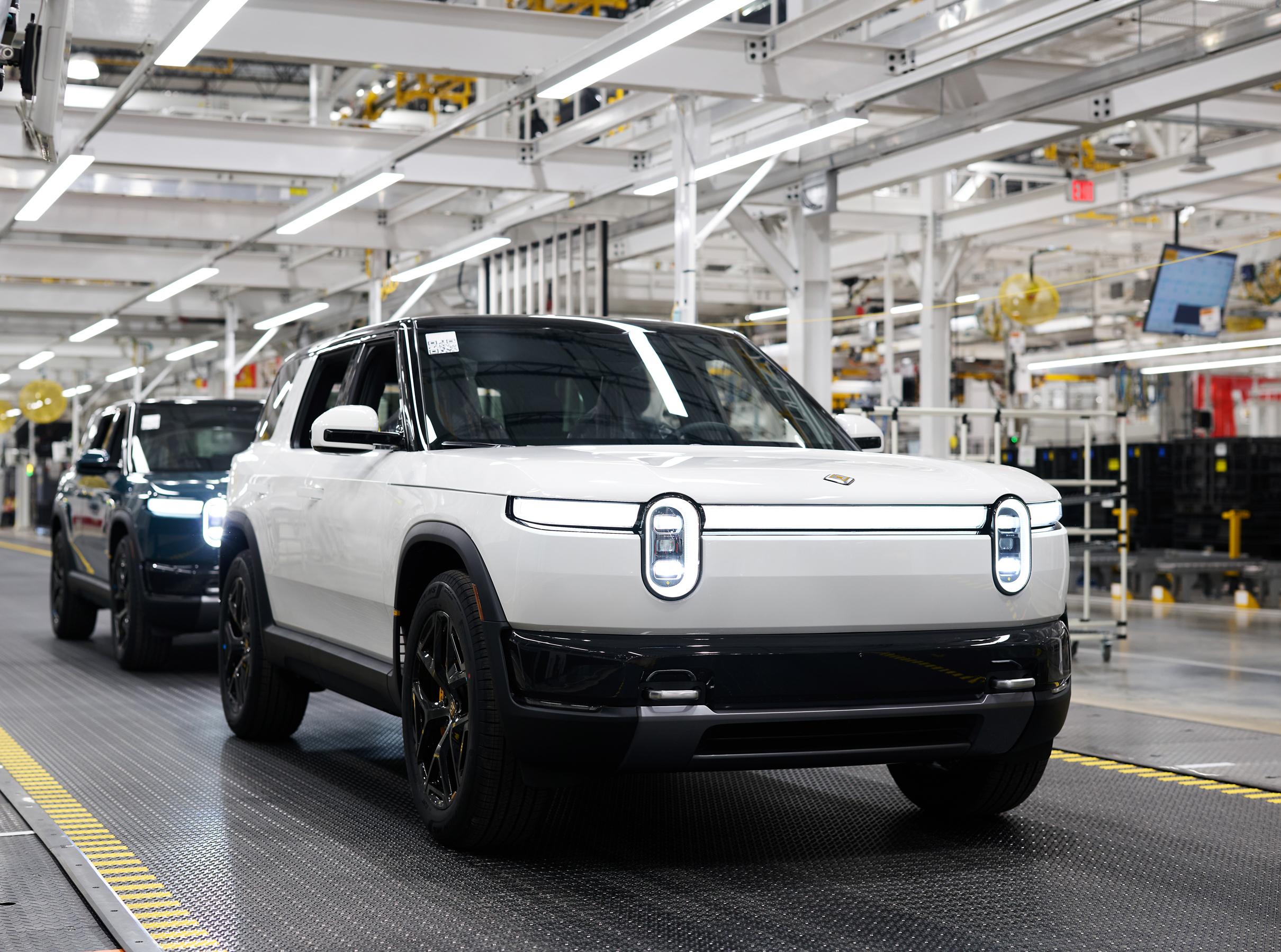 Two Rivian R2's with headlights on are displayed, the first is white and the one behind is blue. Two Rivian R2's with headlights on are displayed, the first is white and the one behind is blue.