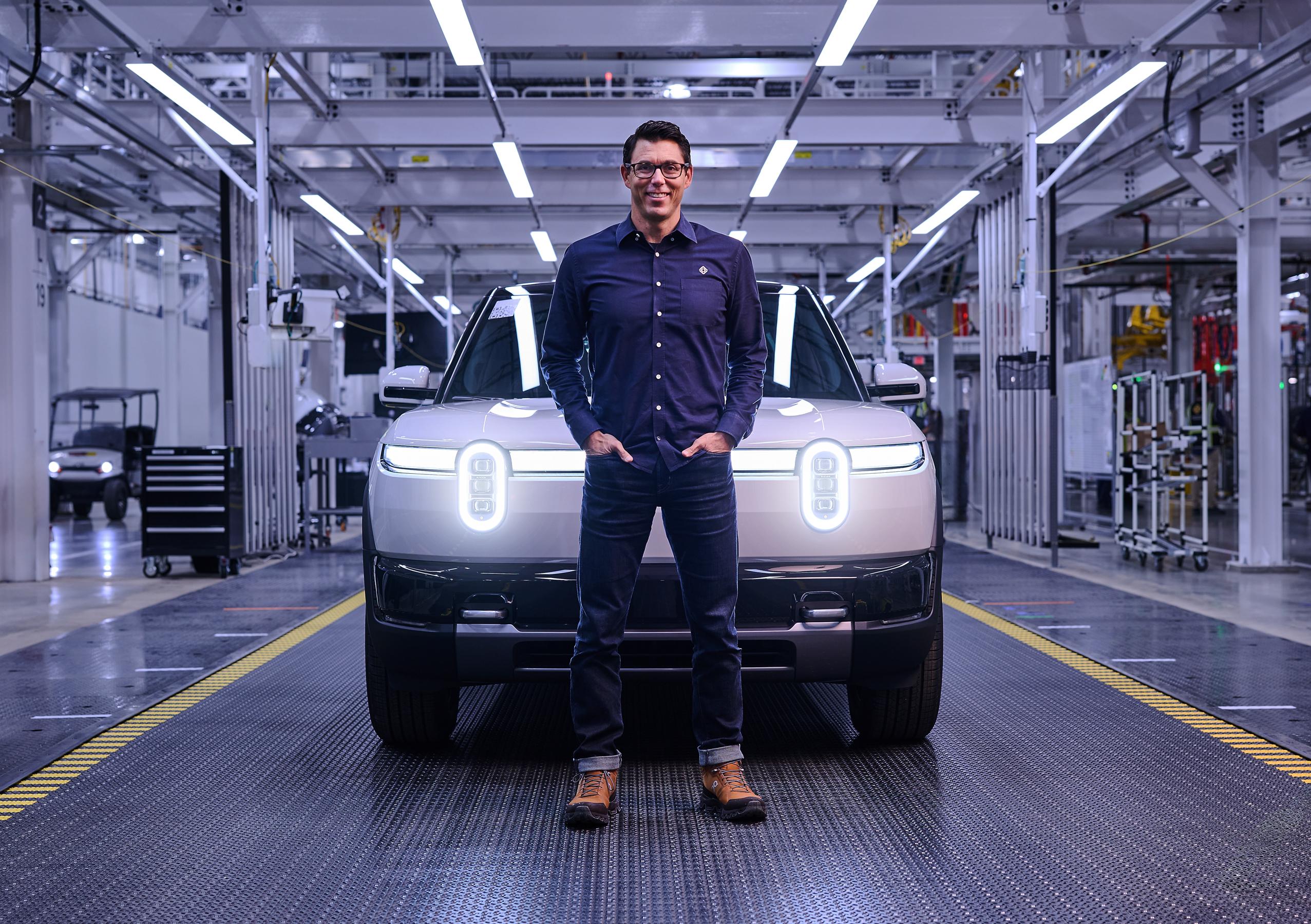 A smiling man wearing jeans, a blue button down shirt, boots and glasses stands in front of a white Rivian R2 with its headlights on. The image is taken in a factory and has a blue cast. A smiling man wearing jeans, a blue button down shirt, boots and glasses stands in front of a white Rivian R2 with its headlights on. The image is taken in a factory and has a blue cast.