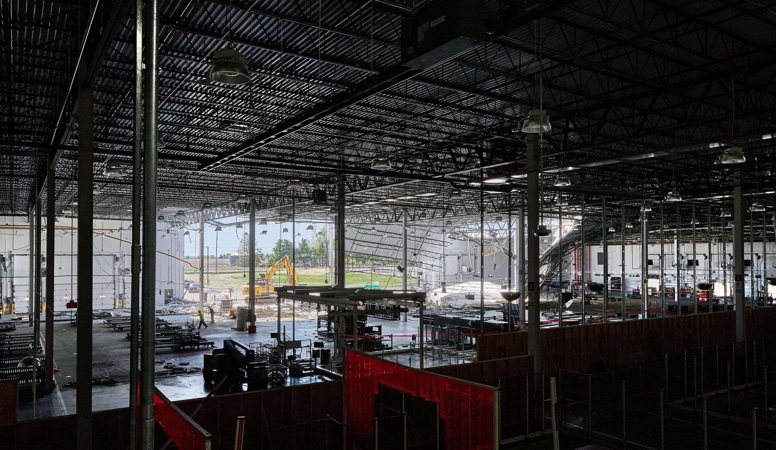 A wide view of a factory interior is mostly dark with a large open wall in the back where you can see to the bright daylight outside. A construction vehicle is in the center and debris can be seen hanging down. A wide view of a factory interior is mostly dark with a large open wall in the back where you can see to the bright daylight outside. A construction vehicle is in the center and debris can be seen hanging down.