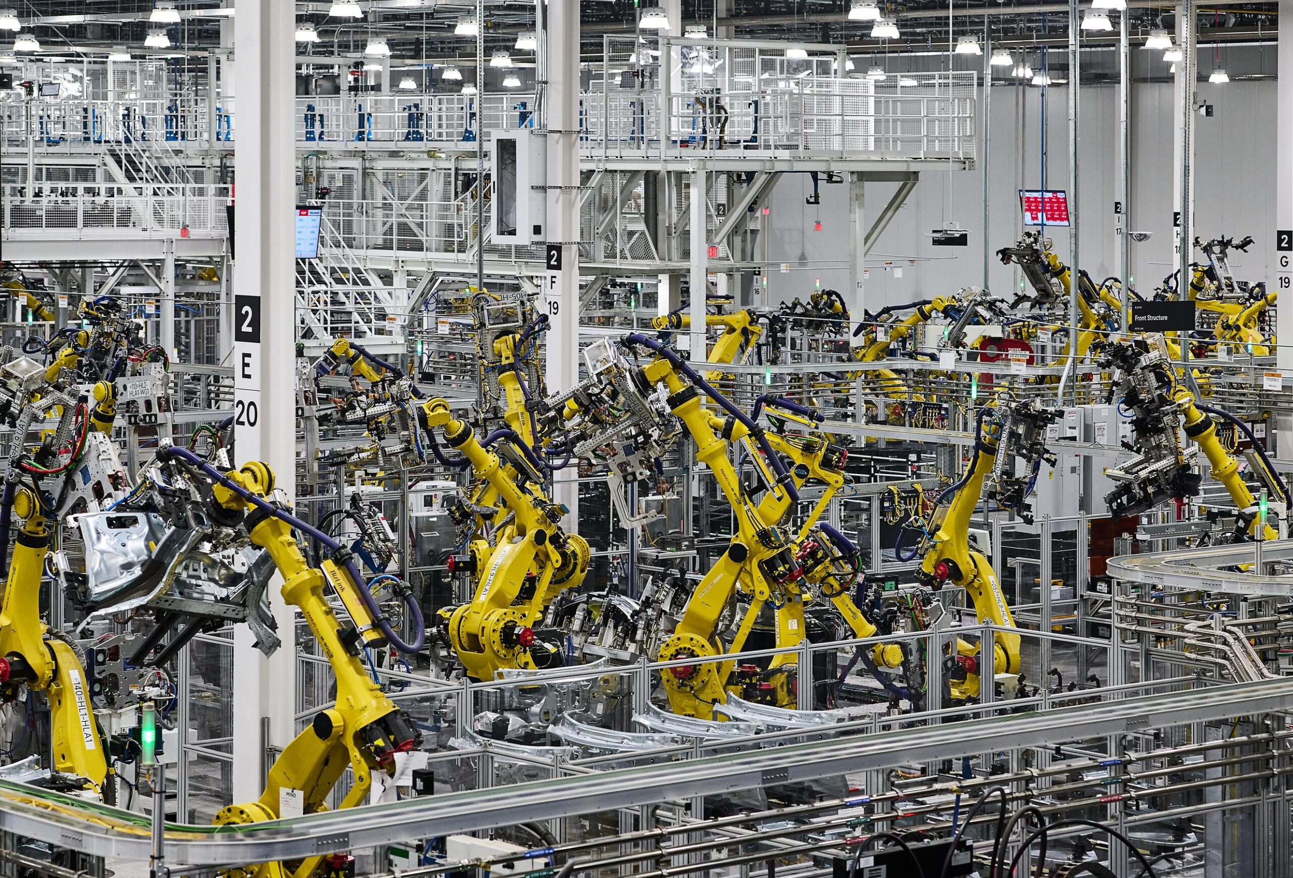 A wide and high view of the factory floor where yellow machine arms are seen lifting and working on unspecified car parts. A wide and high view of the factory floor where yellow machine arms are seen lifting and working on unspecified car parts.