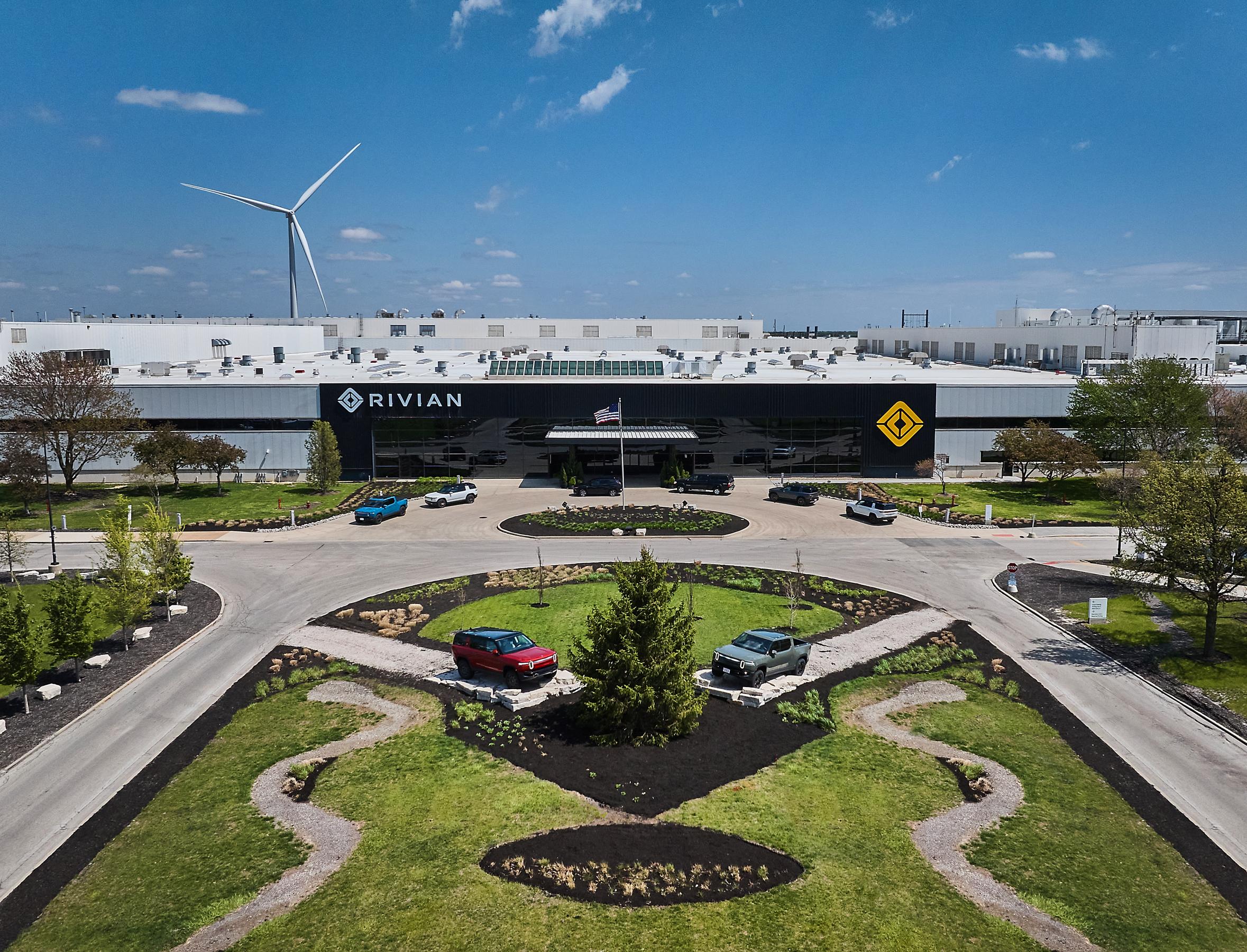 A drone shot of the entrance of the Rivian factory. There's a manicured grassy area with two vehicles displayed along the road leading to the entrance of the enormous Rivian building. It's a bright sunny day with a blue sky overhead. A drone shot of the entrance of the Rivian factory. There's a manicured grassy area with two vehicles displayed along the road leading to the entrance of the enormous Rivian building. It's a bright sunny day with a blue sky overhead.