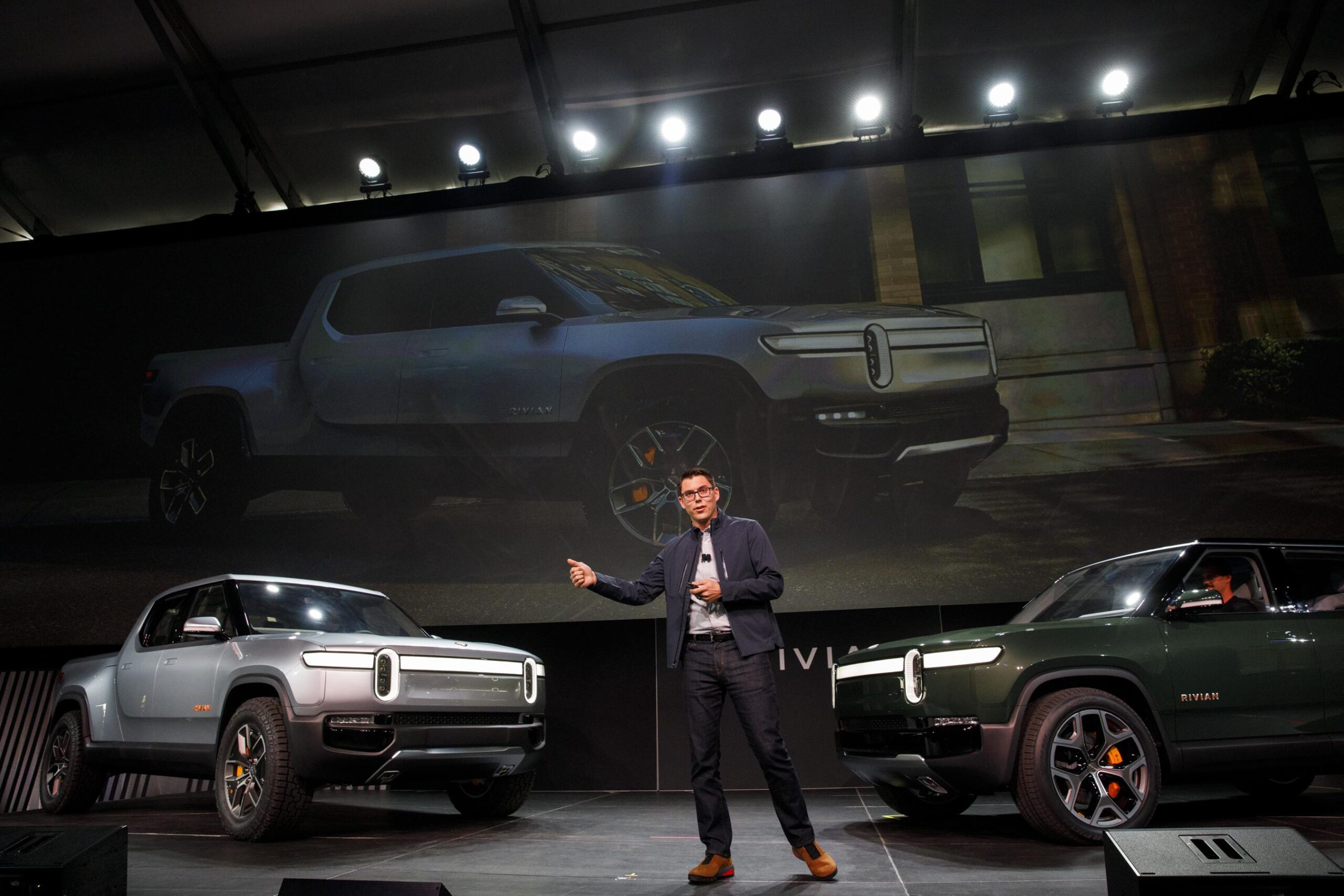 A man with glasses stands on stage with two Rivian vehicles facing him. A man with glasses stands on stage with two Rivian vehicles facing him.