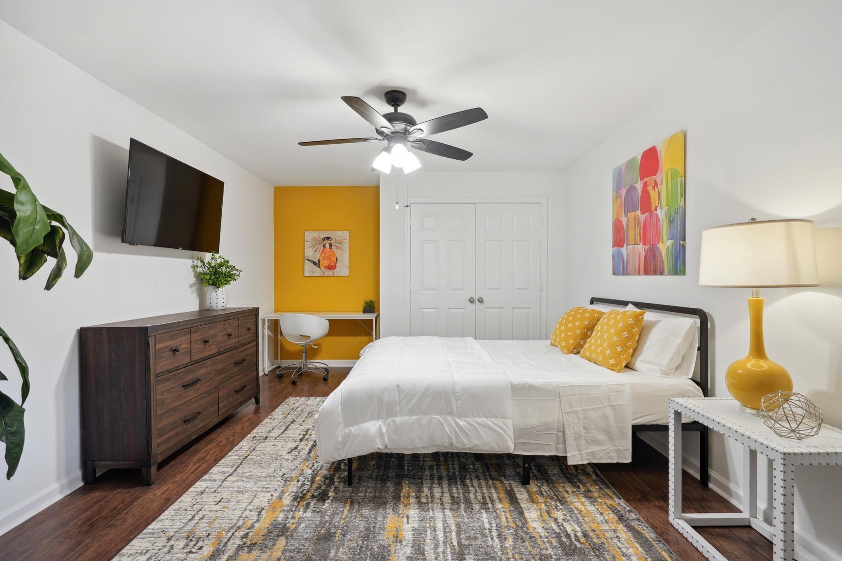 A small bedroom with queen bed, ceiling fan, and a TV hanging above a dresser.
