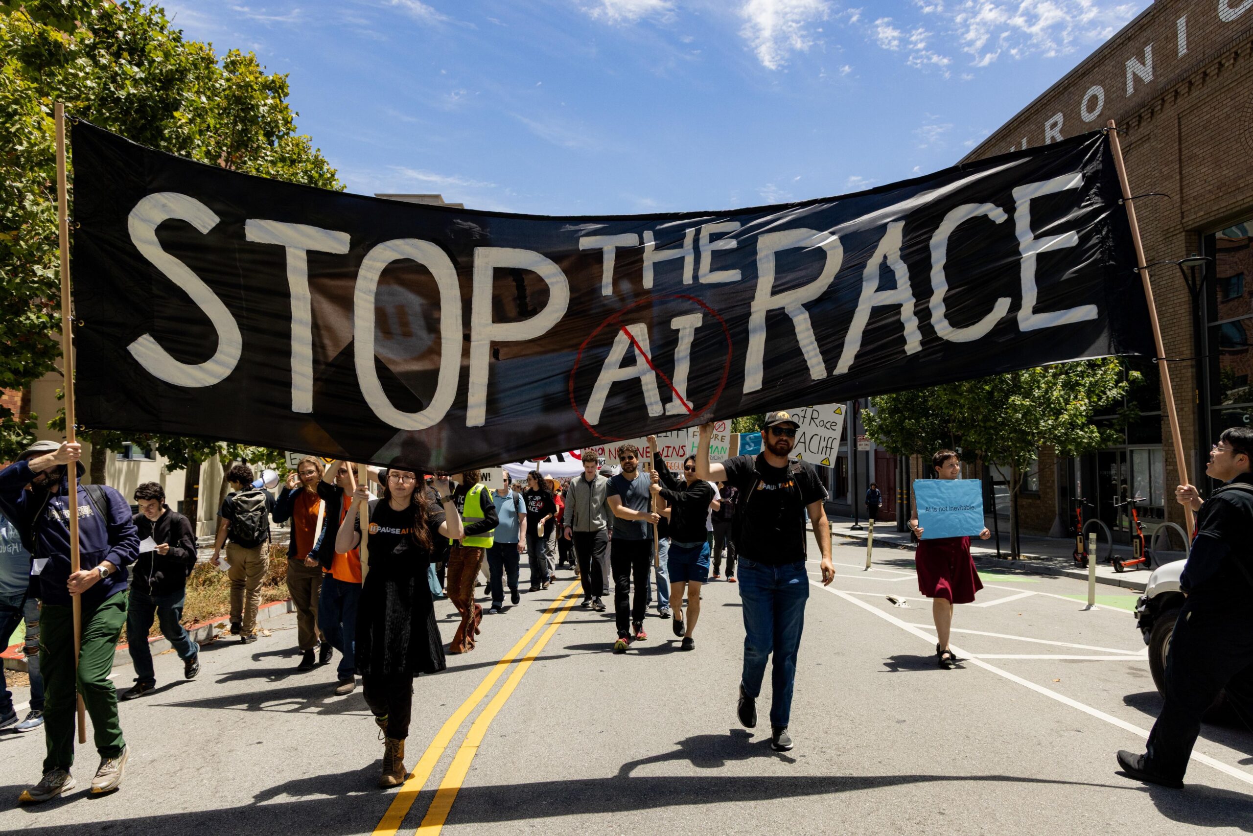 Protesters march down a sunny city street beneath a large black banner reading “STOP THE AI RACE,” with the letters “AI” crossed out in red.