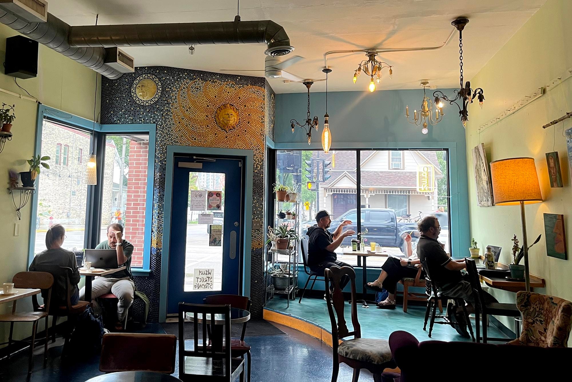 a blue, green, and yellow cafe interior with mosaic art and eclectic hanging lights. Several people are eating and working at small tables.