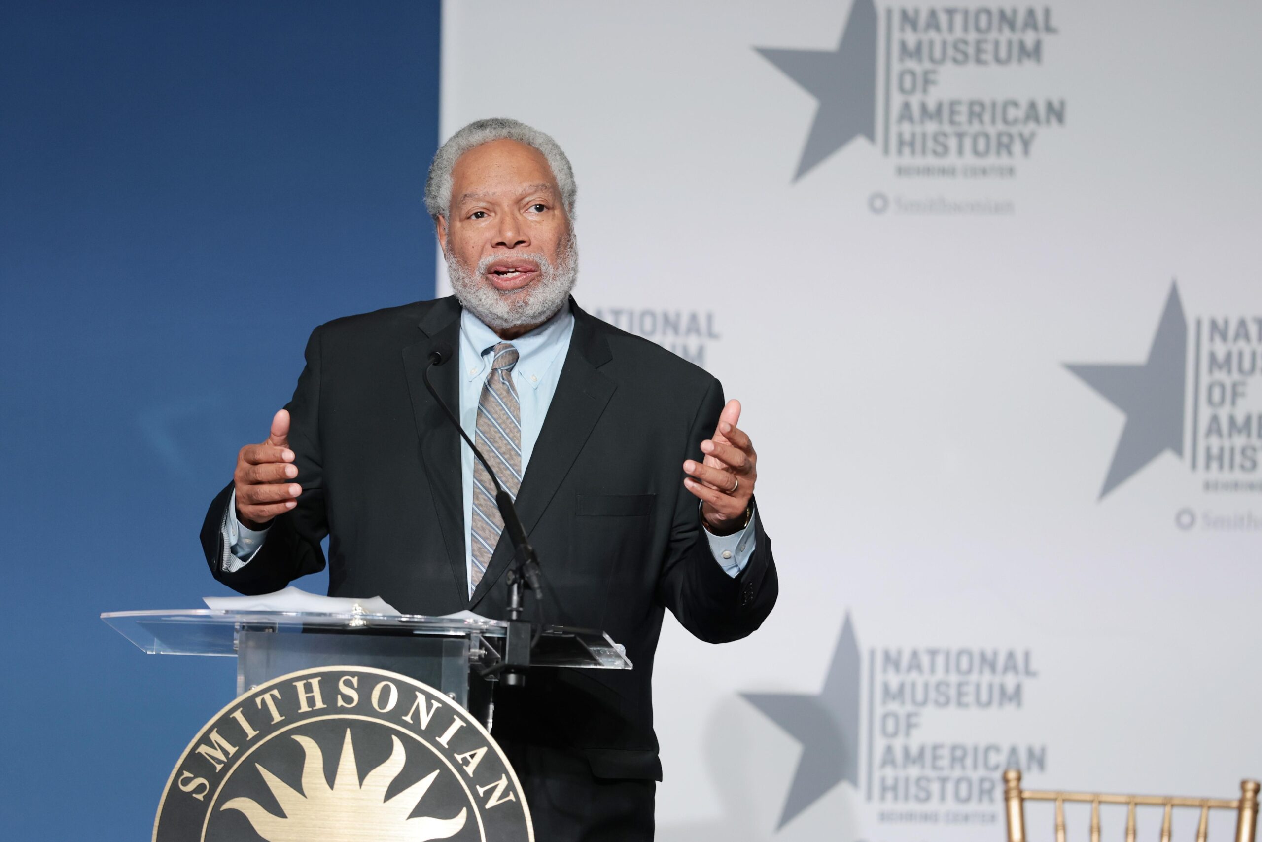 Lonnie Bunch, a bearded, white-haired man wearing a suit, gestures while speaking from behind a podium at an event at the Smithsonian National Museum of American History.