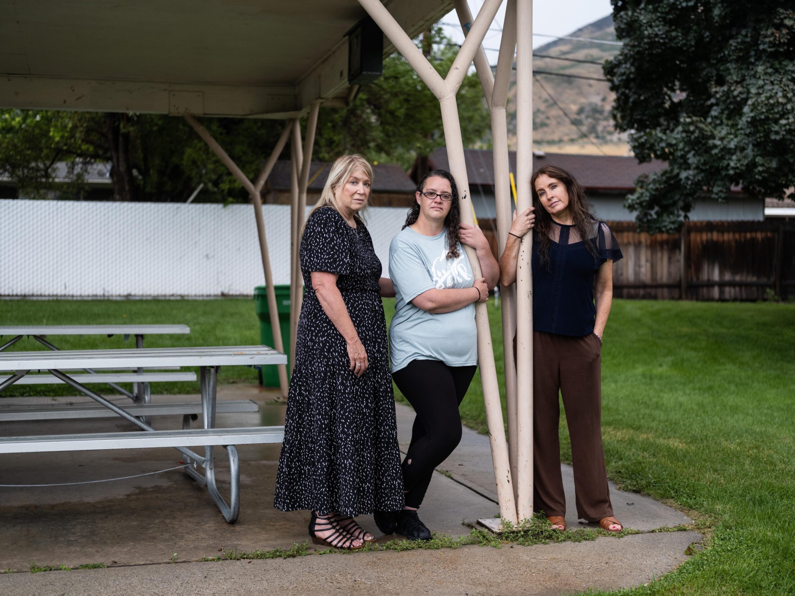 Three women stand side by side beneath a picnic awning in a park. 