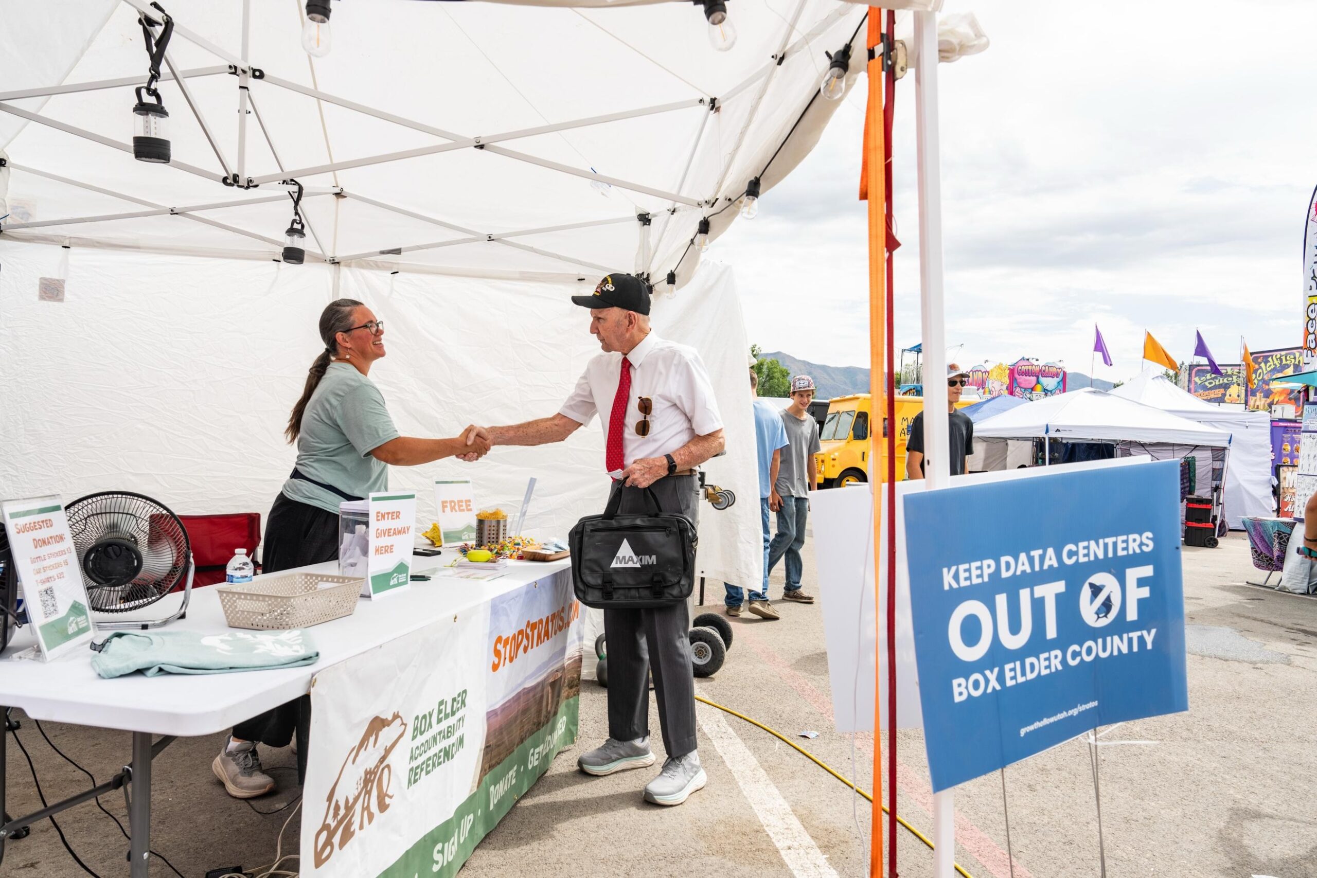 A man and a woman shaking hands beneath a pop-up tent with a sign in the foreground that reads "keep data centers out of Box Elder County."