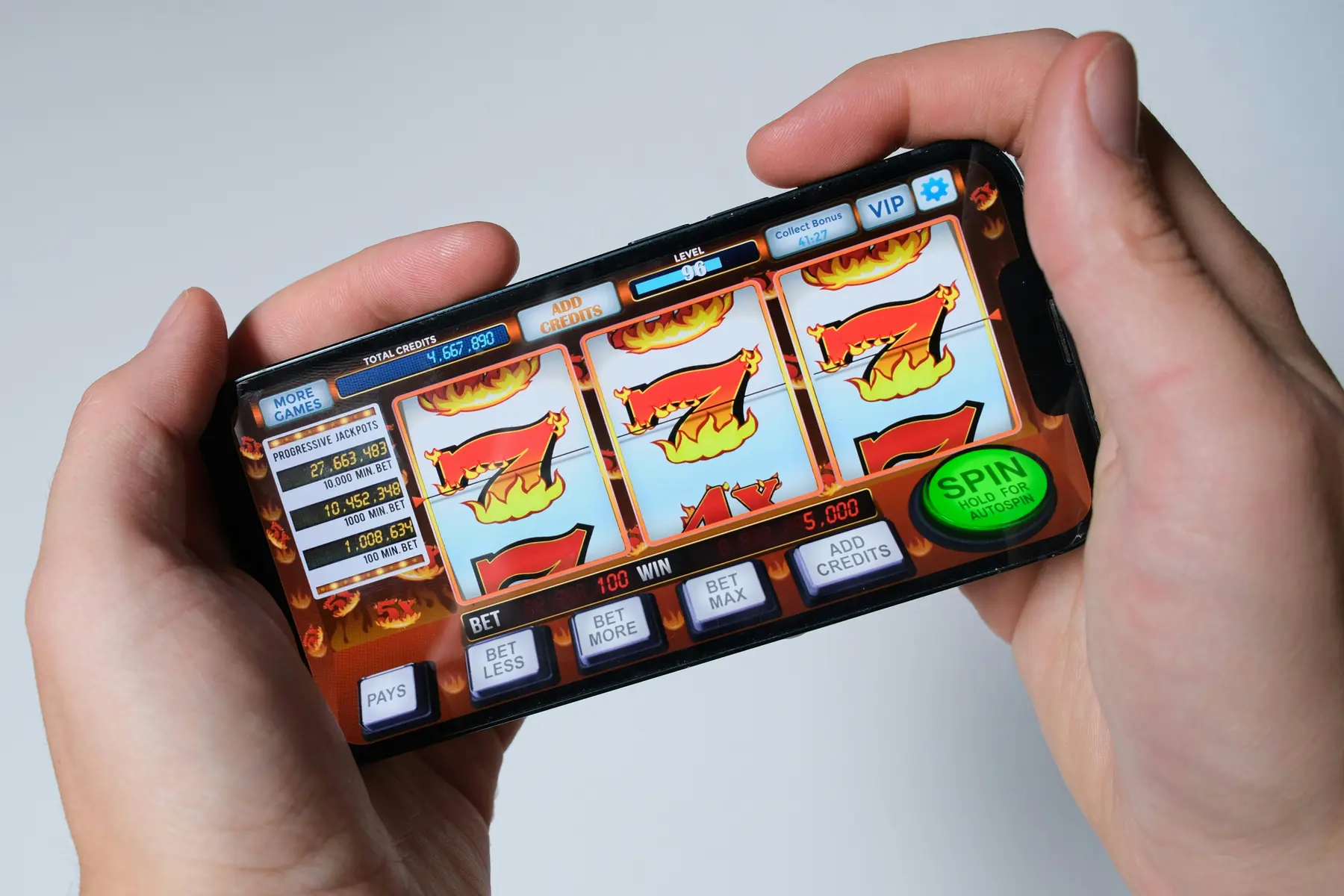 A person holding a smartphone displaying a mobile slot machine game with spinning reels and a spin button.